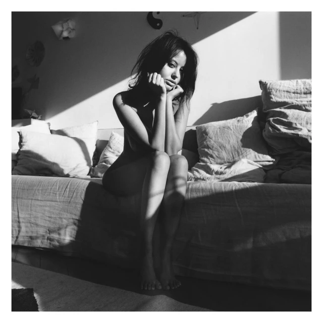 Monochrome image of a young adult woman with dark hair, sitting on a daybed with pillows, backlit by sunlight coming through a window.