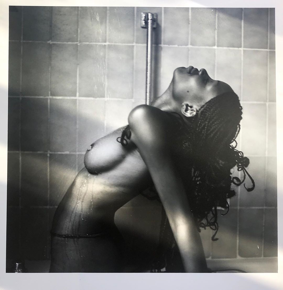 Striking black-and-white photo of a nude woman with braids, back arched, facing up toward a showerhead in a tiled shower.