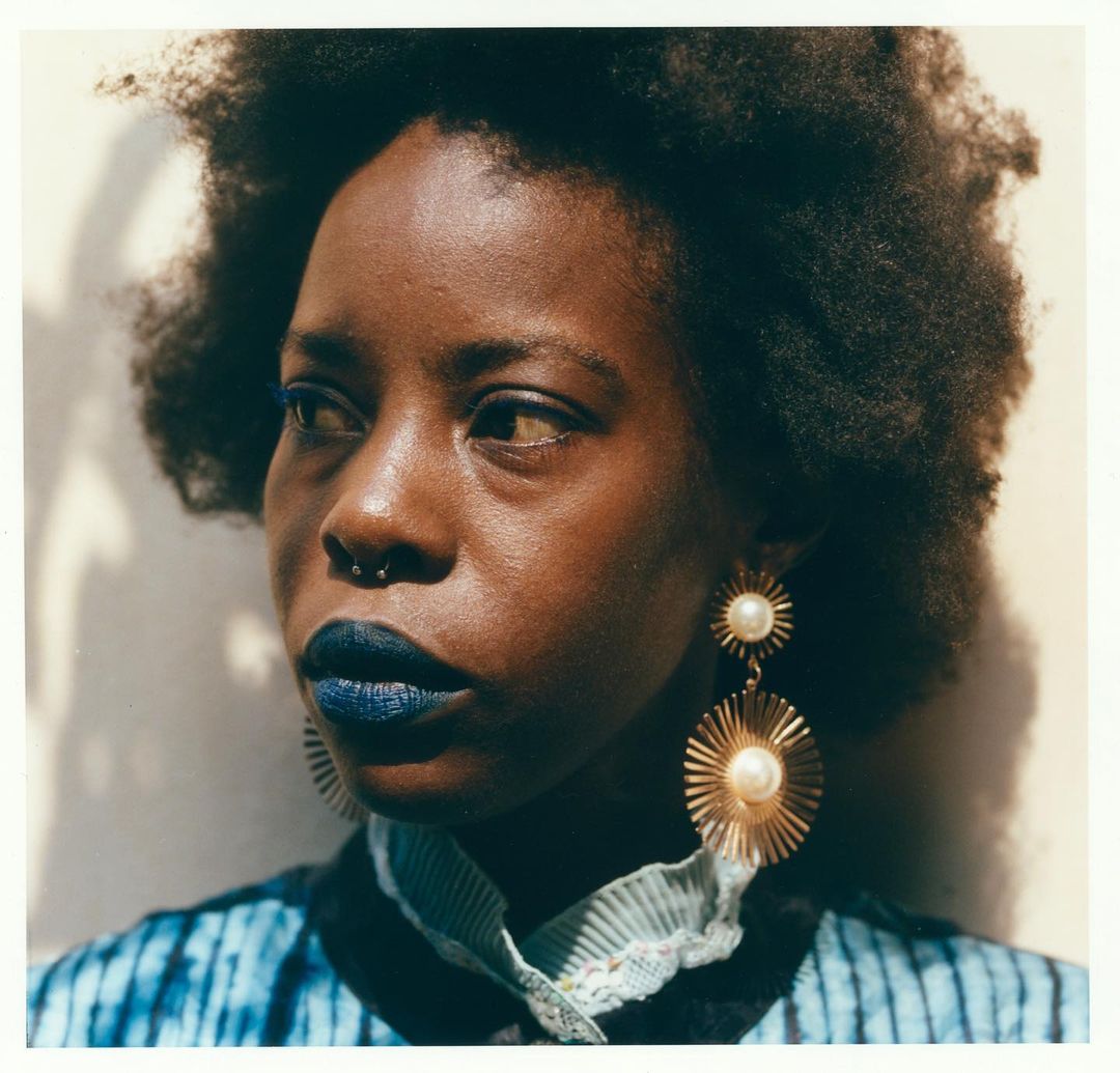 Close-up portrait of a Black woman with an afro wearing statement pearl earrings and blue lipstick, looking off to the side.