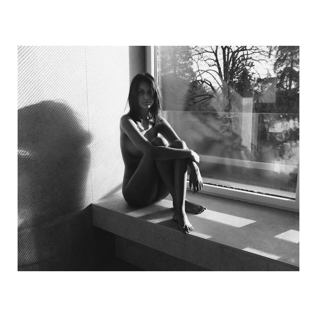 Black and white portrait of a nude woman sitting by a window with trees outside, casting shadows, creating a contemplative scene.