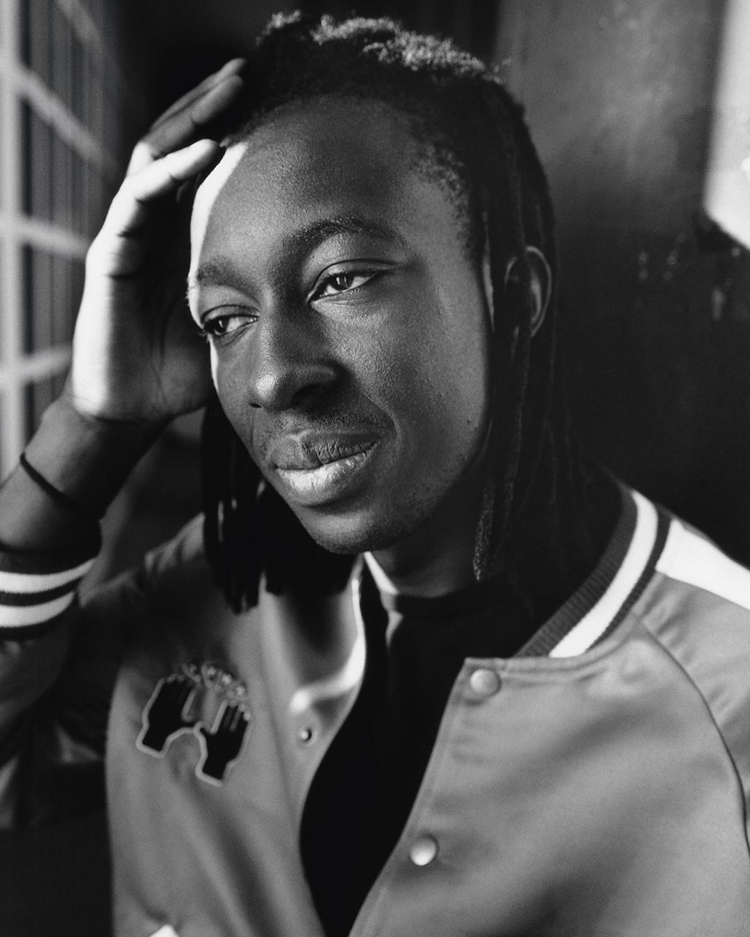 Monochromatic portrait of a Black man with locs, wearing a varsity jacket, looking slightly off-camera, with his hand on his head.