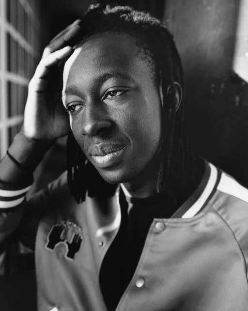 Monochromatic portrait of a Black man with locs, wearing a varsity jacket, looking slightly off-camera, with his hand on his head.