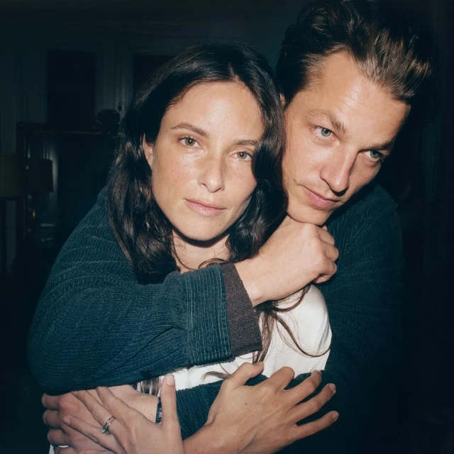 Portrait of a couple indoors, woman with dark hair looks at the camera while embraced by a man with short brown hair.