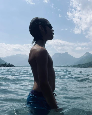Profile of a young boy wearing sunglasses standing waist-deep in a lake with distant mountain peaks under a cloudy sky.
