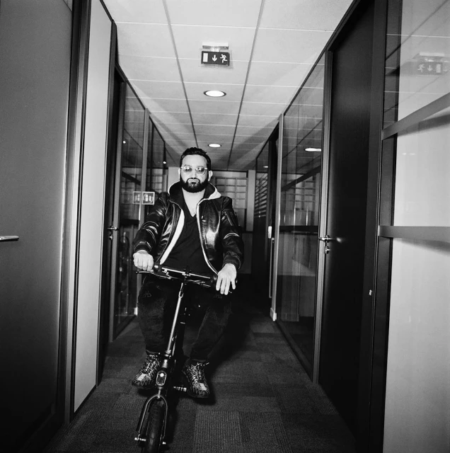 Black and white shot of a man riding an electric scooter down an office hallway, wearing a jacket and sunglasses.