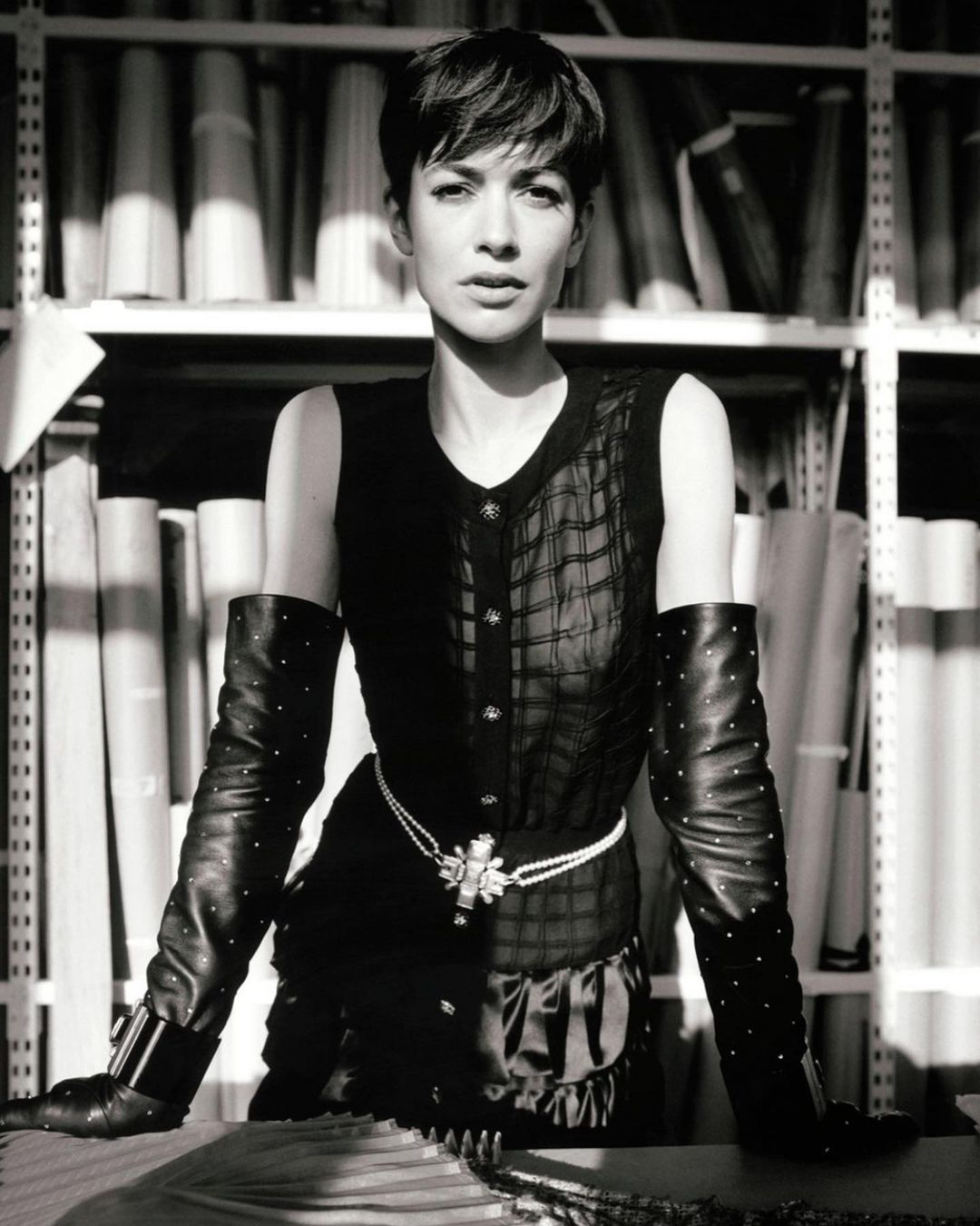 Black-and-white portrait of a short-haired woman in a sleeveless top, long gloves, and a pearl belt, posing in an indoor setting.