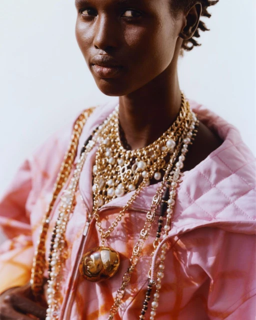 Close-up of a dark-skinned adult female model wearing an open pink quilted jacket and layered necklaces, against a bright background.