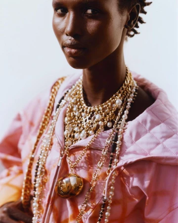 Close-up portrait of a woman wearing layered gold chain necklaces and a pink quilted jacket, photographed by Paul Schmidt.