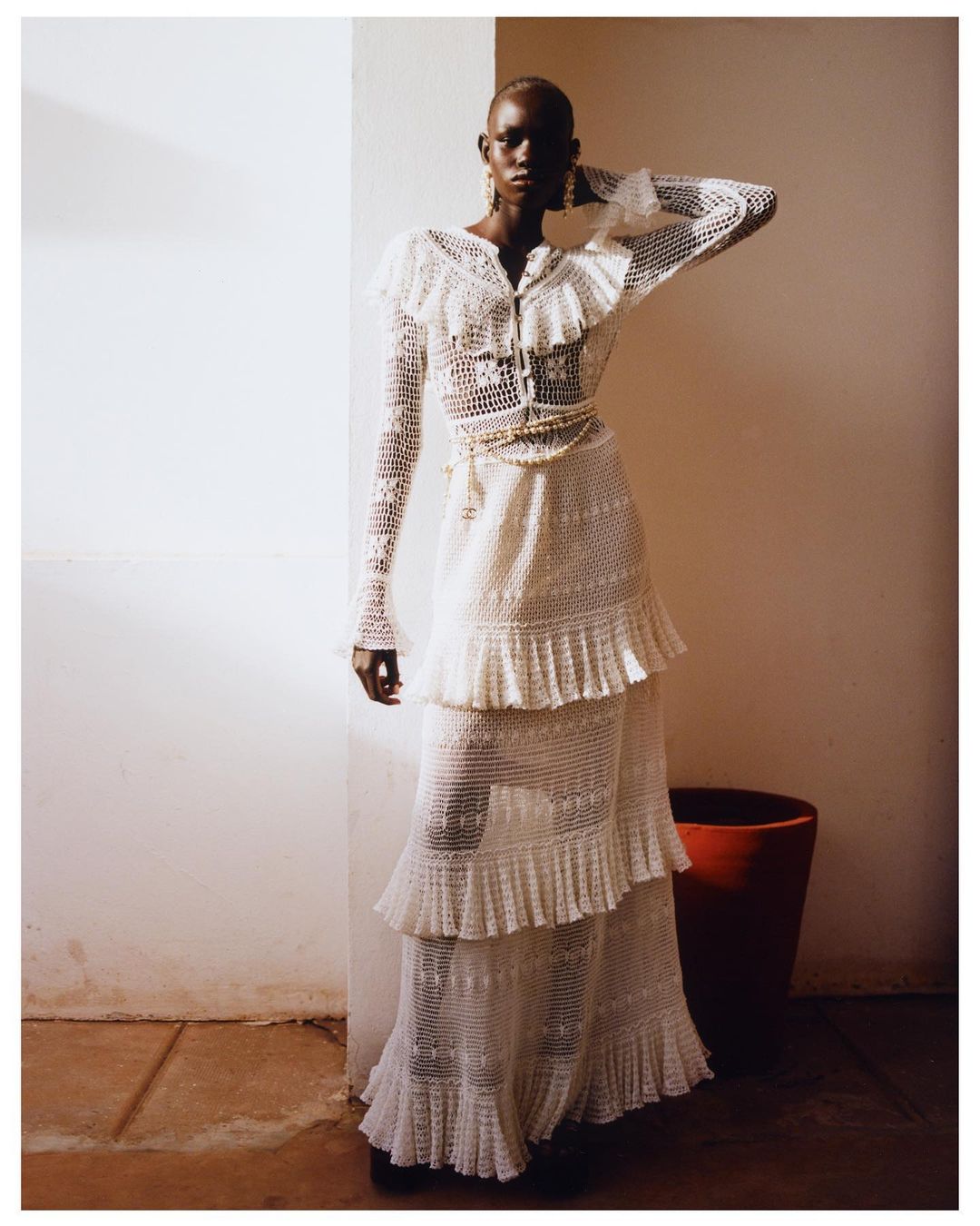 A Black female model poses indoors wearing a long, cream-colored, tiered crochet dress with ruffle details and drop earrings in a fashion shot.