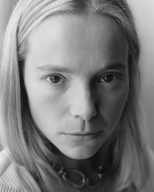 Monochrome portrait of a fair-skinned woman with long blonde hair and a neutral expression, wearing a linked silver necklace.