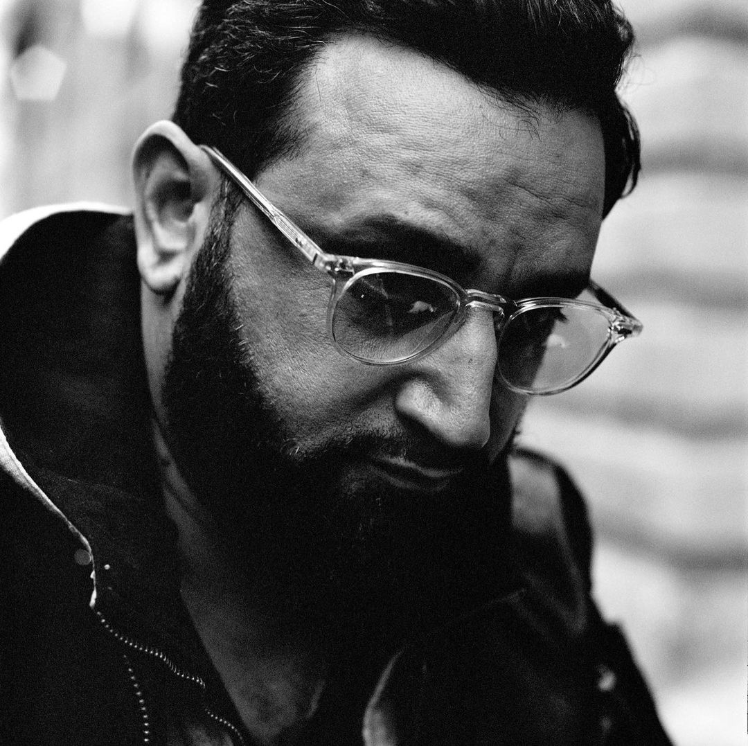 Close-up monochrome portrait of a bearded man wearing clear-framed glasses and a dark jacket by photographer Paul Schmidt.