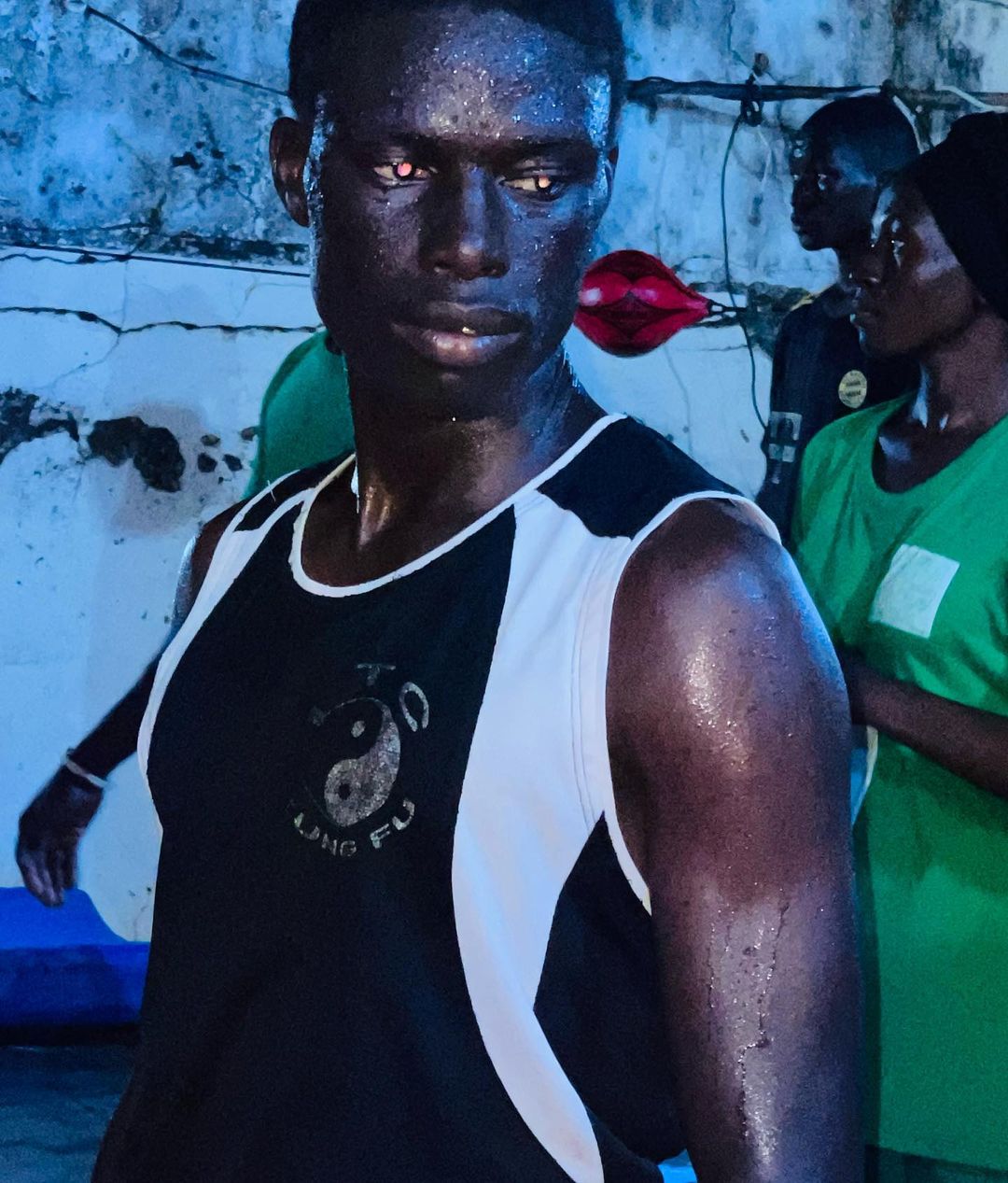 Sweaty adult male athlete wearing a Kung Fu tank top in a gym setting, with other people and boxing equipment in the background.