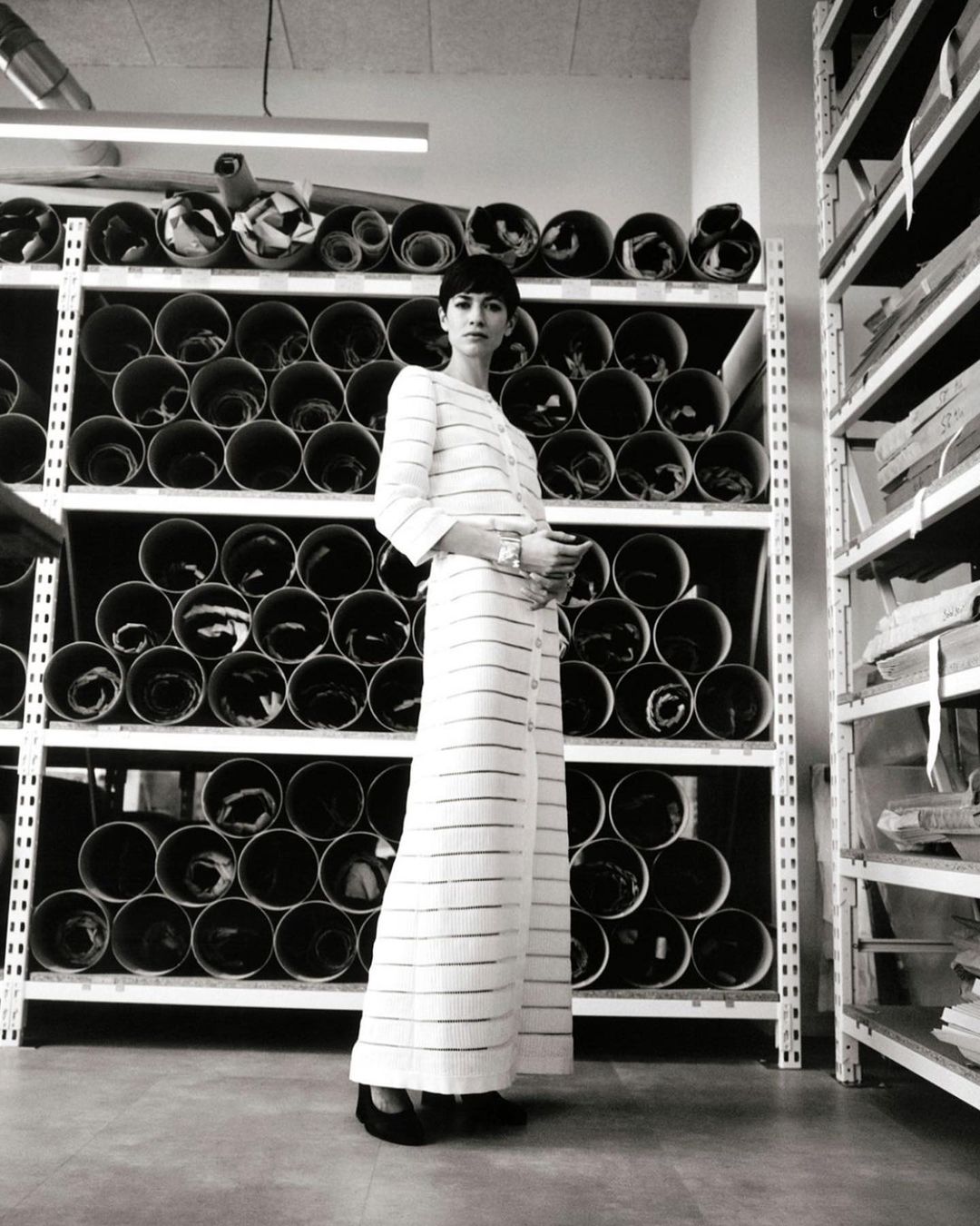A woman in a striped pantsuit stands in front of shelves of rolled textiles, photographed in a warehouse-like space with neutral tones.