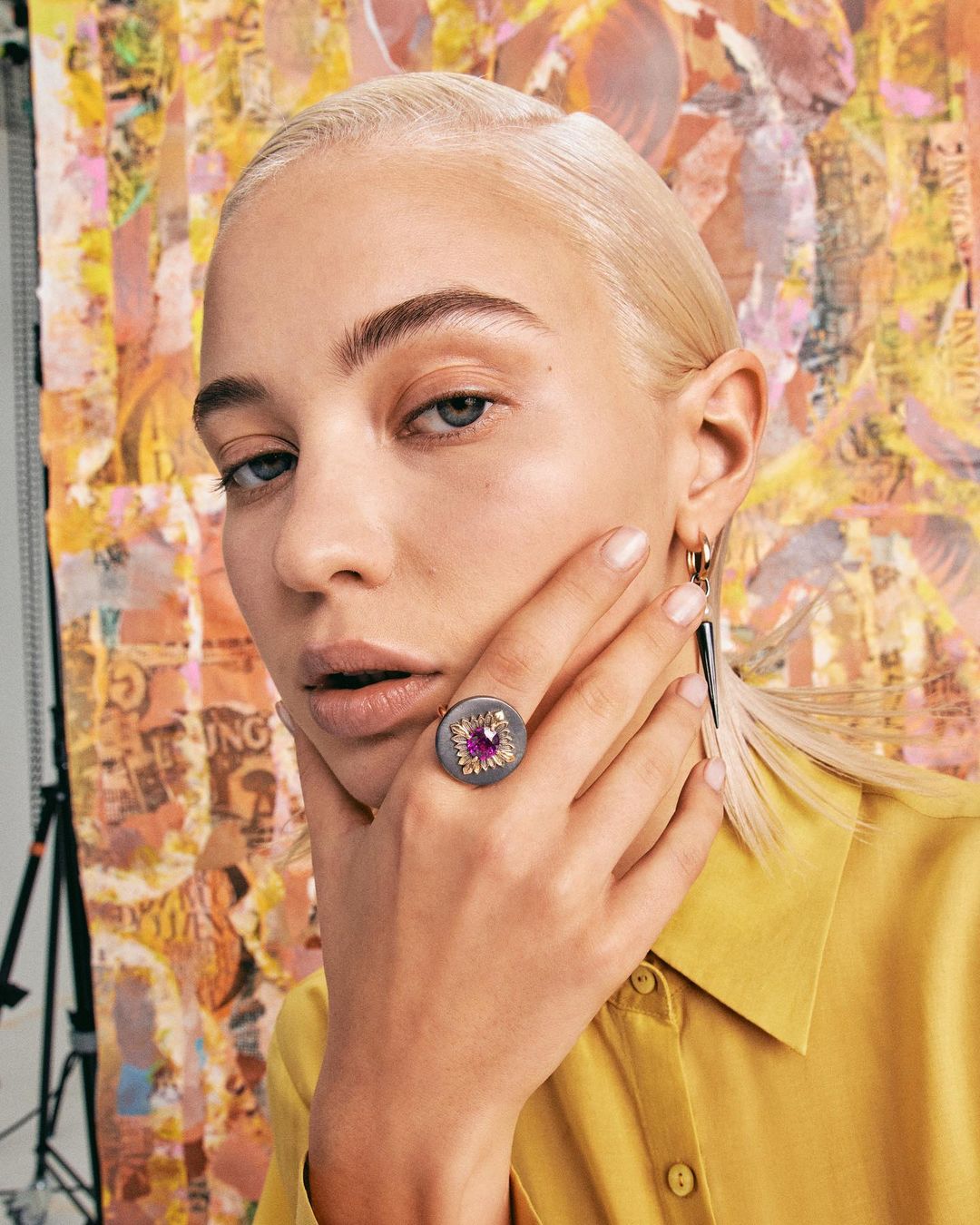 Close-up portrait of an adult woman with platinum hair and yellow top wearing a large ring against an abstract patterned background.
