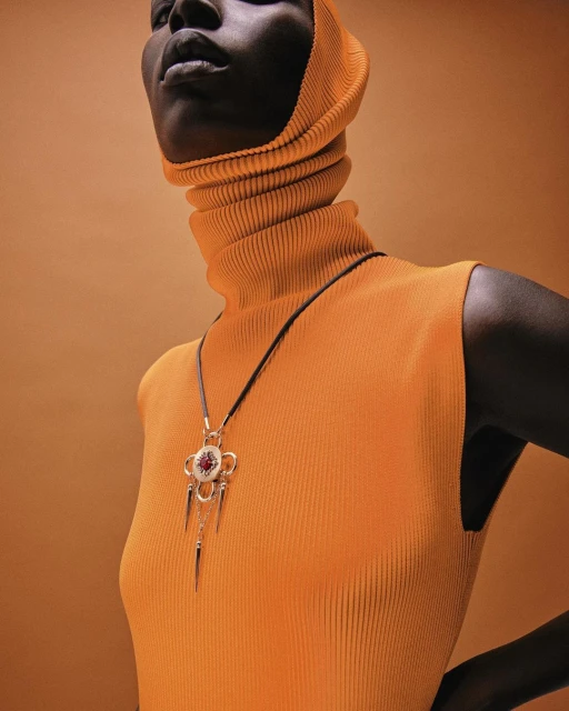 Close-up of an African-American female fashion model with a gold necklace and an orange ribbed turtleneck and hood against an orange background.