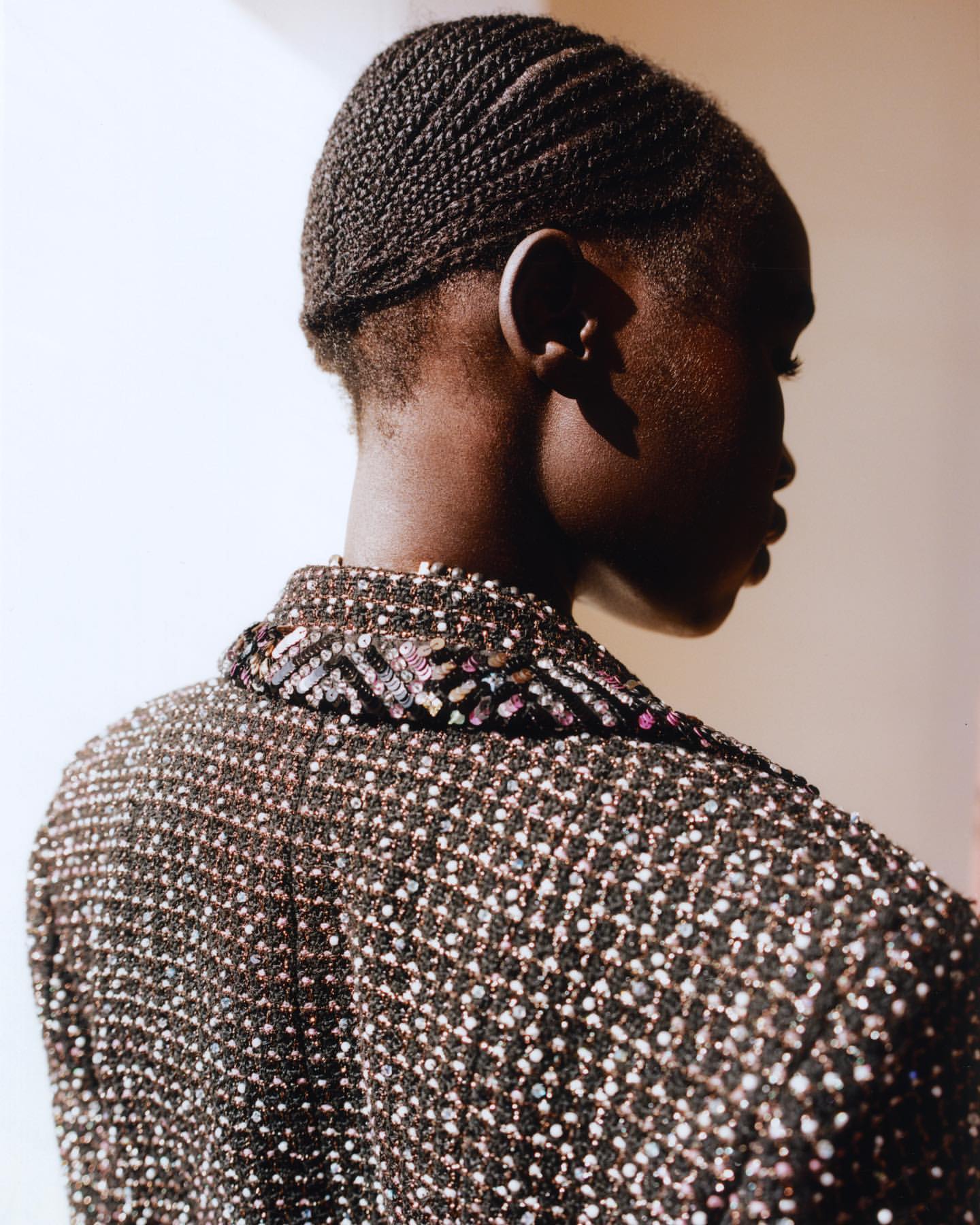 Woman with dark braided hair, seen from the side, wearing a sequined jacket with an ornate collar, posing against a neutral background.