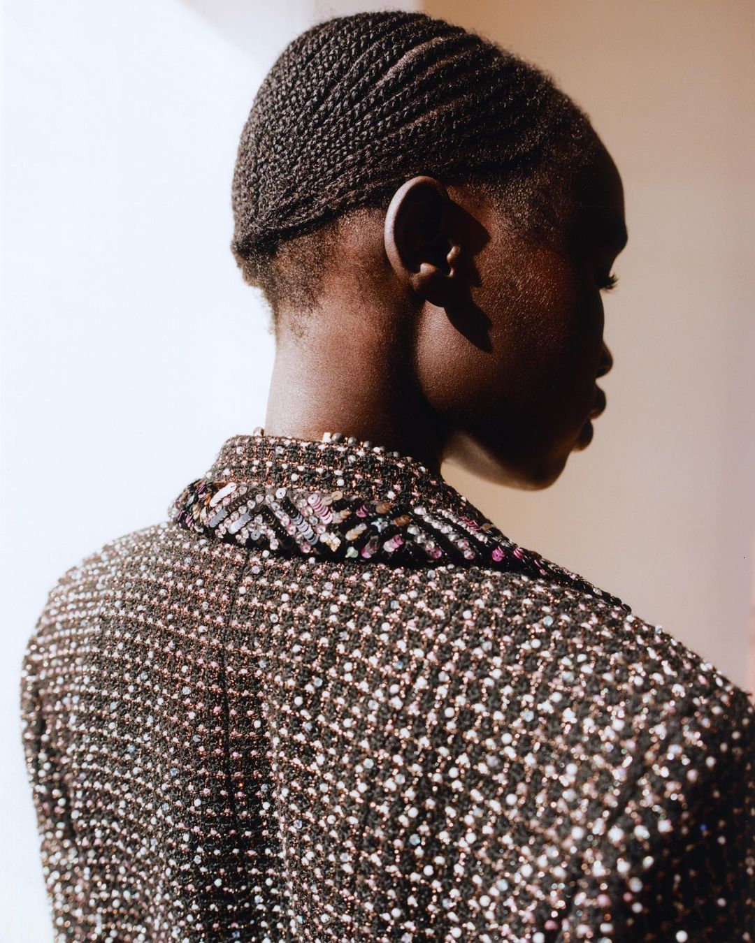 Back view of African-American fashion model with braided hair wearing a sequined blazer in a studio with soft lighting.
