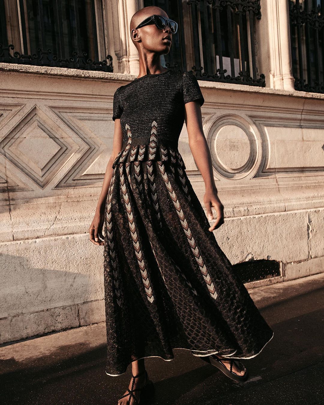 An African-American female adult model wears a black dress on a city sidewalk near a detailed stone wall in natural lighting.