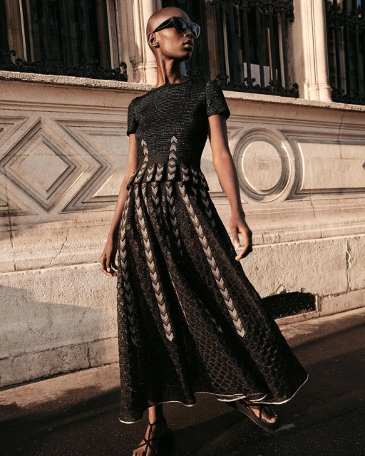 An African-American female adult model wears a black dress on a city sidewalk near a detailed stone wall in natural lighting.