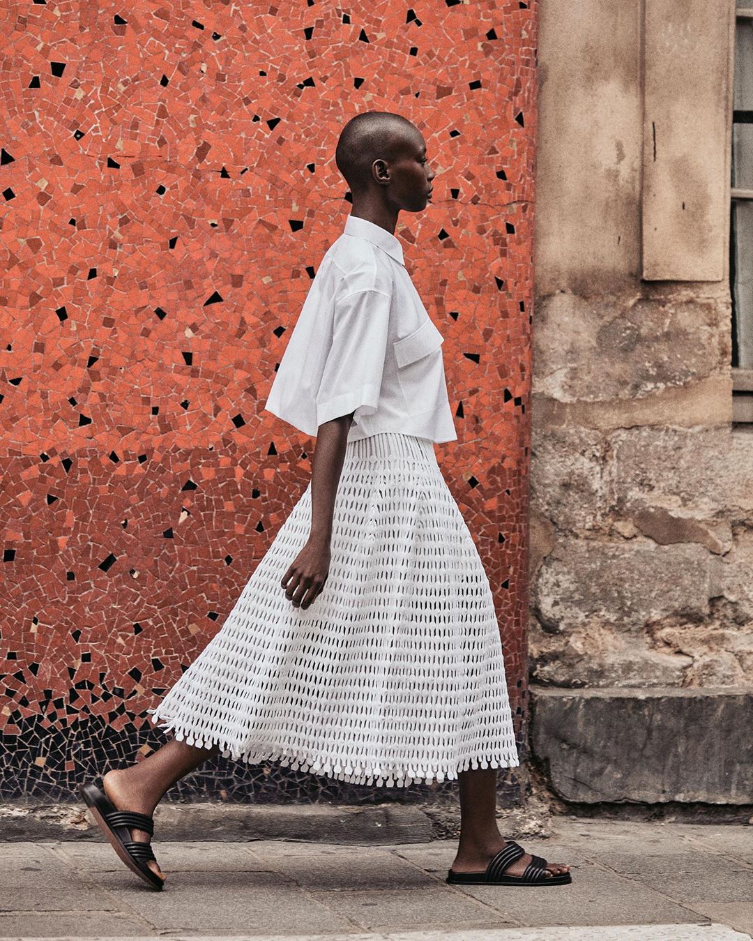 Woman in white shirt and skirt walks past a mosaic wall on a city street, showcasing fashion apparel and urban style.