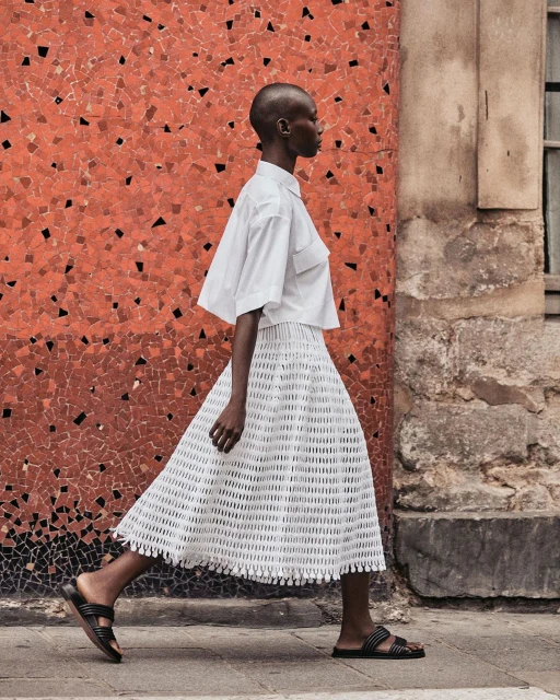 Woman in white shirt and skirt walks past a mosaic wall on a city street, showcasing fashion apparel and urban style.