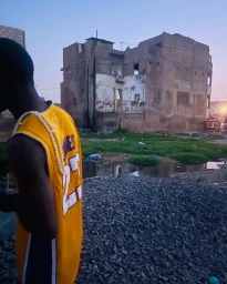 Black man wearing a yellow basketball jersey stands in front of a gravel area and an unfinished brick building.