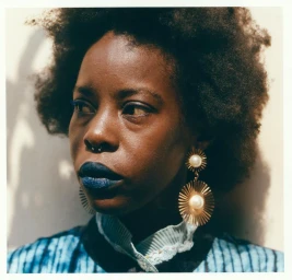 Close-up portrait of a Black woman with dark blue lipstick, septum ring, and large gold earrings against a light wall