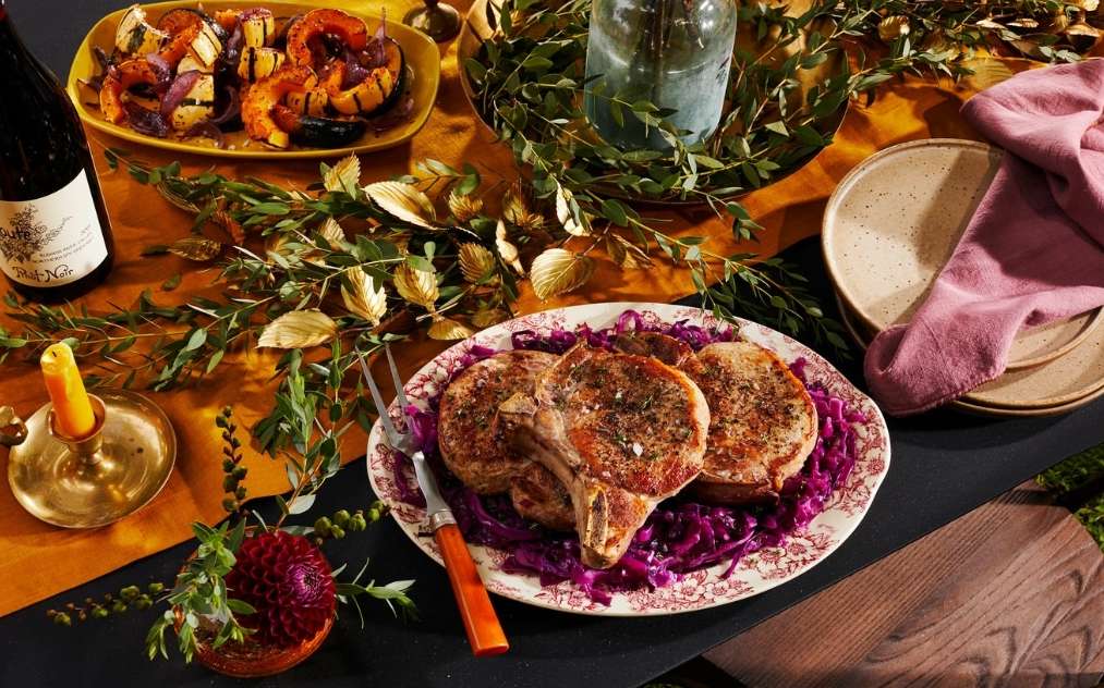 Overhead shot of an elegant autumn dinner with pork chops on red cabbage, roasted squash, wine, candle, and foliage on a table.