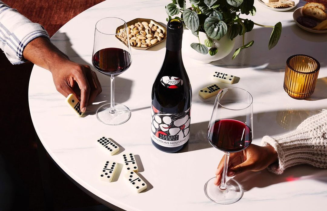 Overhead shot of a wine bottle and two wine glasses filled with red wine, surrounded by dominoes, pistachios, and hands on a white marble table.