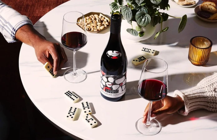 Overhead shot of a wine bottle and two wine glasses filled with red wine, surrounded by dominoes, pistachios, and hands on a white marble table.