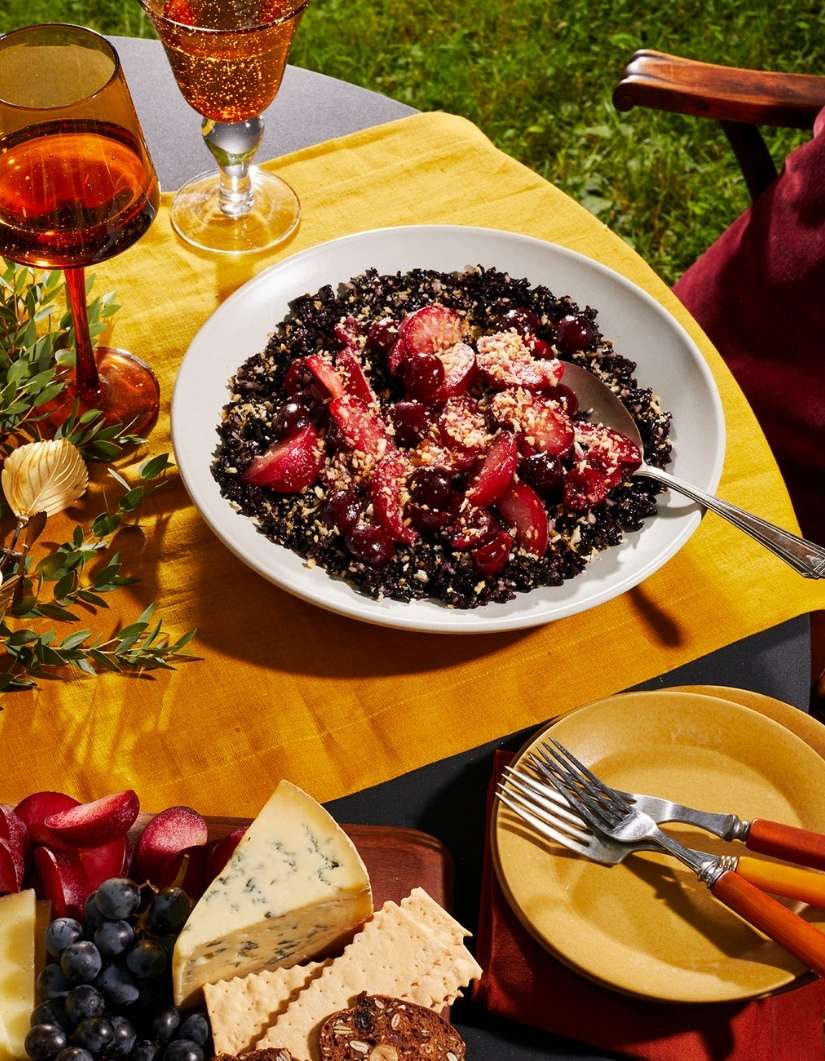 Elevated shot of black rice and plums on a white plate, accompanied by wine, cheese, grapes, and crackers, set outdoors on a yellow cloth.