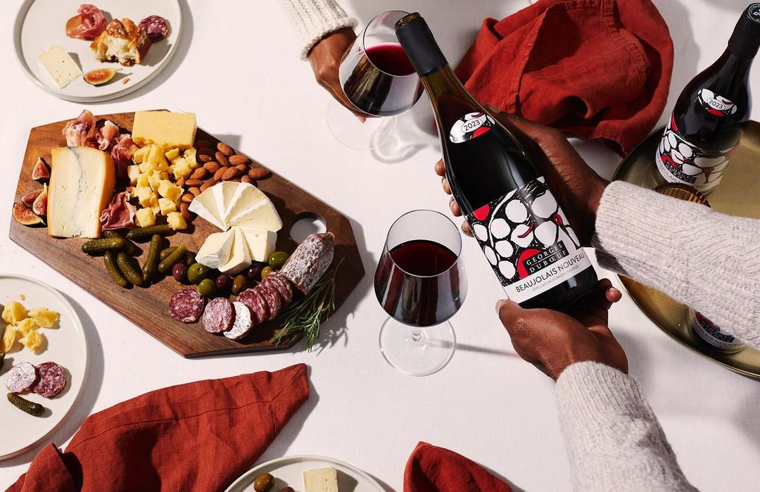 Overhead shot of two people holding a wine bottle and glasses, accompanied by a charcuterie board with assorted cheeses, meats, and olives.