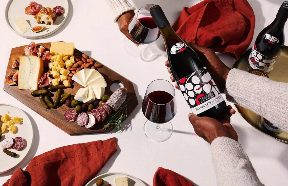 Overhead shot of two people holding a wine bottle and glasses, accompanied by a charcuterie board with assorted cheeses, meats, and olives.