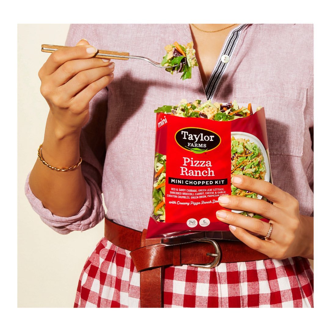 Woman holds Taylor Farms Pizza Ranch salad kit and fork with salad. The ingredients include cabbage, carrots, broccoli, and lettuce.