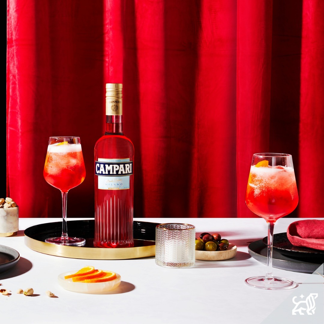 Campari bottle and cocktail glasses on a white table with red curtains background, showcasing a vibrant and elegant drink presentation.