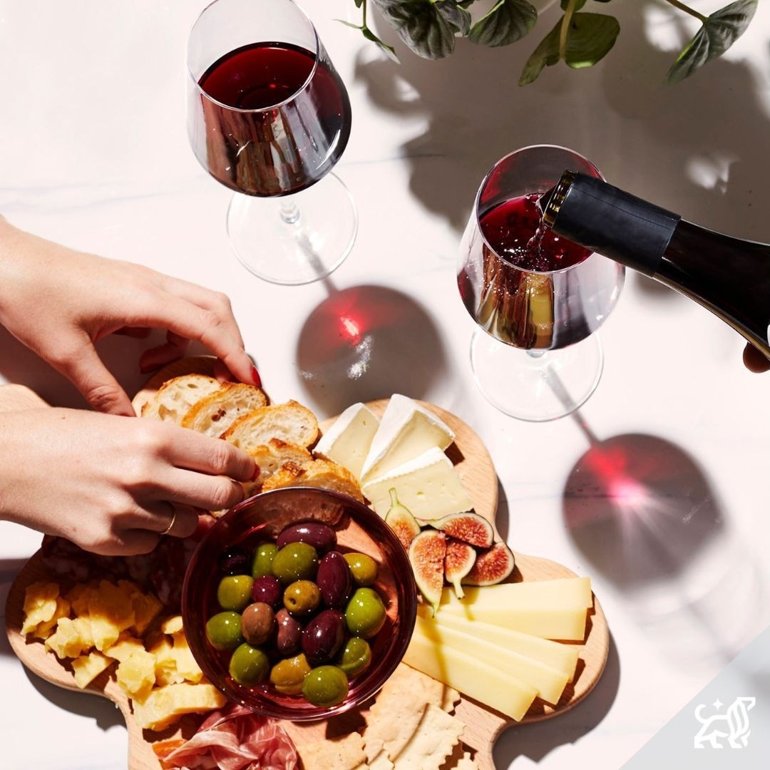 Overhead shot of two wine glasses with red wine being poured alongside a charcuterie board featuring assorted cheeses and bread.