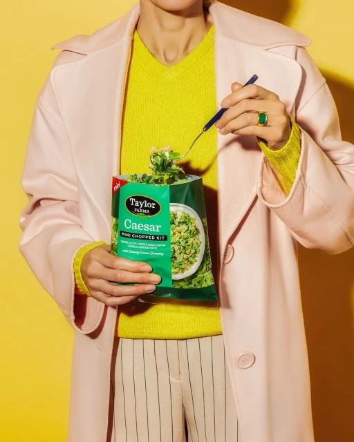 Woman in pastel coat holds Caesar salad kit with fork, showcasing food and fashion against a yellow background in an indoor studio.