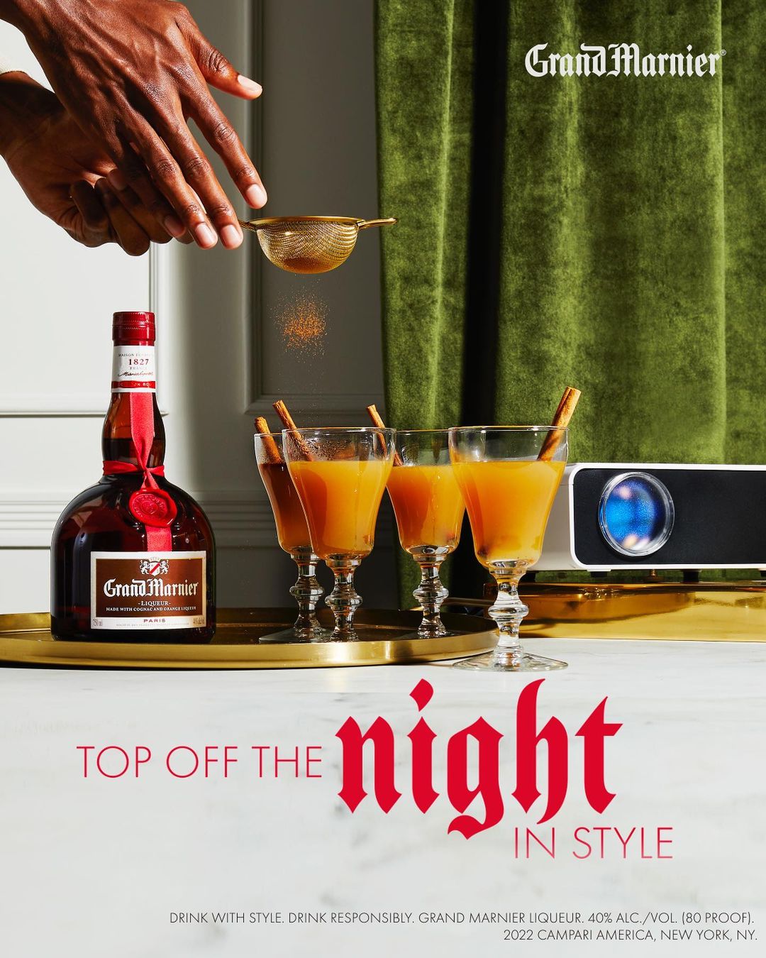 Grand Marnier liqueur bottle with cocktails and an arm sifting spices against a green backdrop. Drinkware on a gold tray.