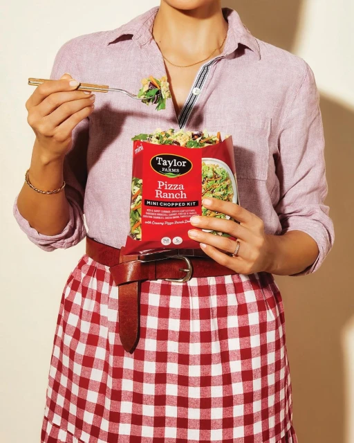 A woman in a red and white checkered skirt and pink linen shirt eats salad with a fork, holding a Taylor Farms Pizza Ranch salad kit.