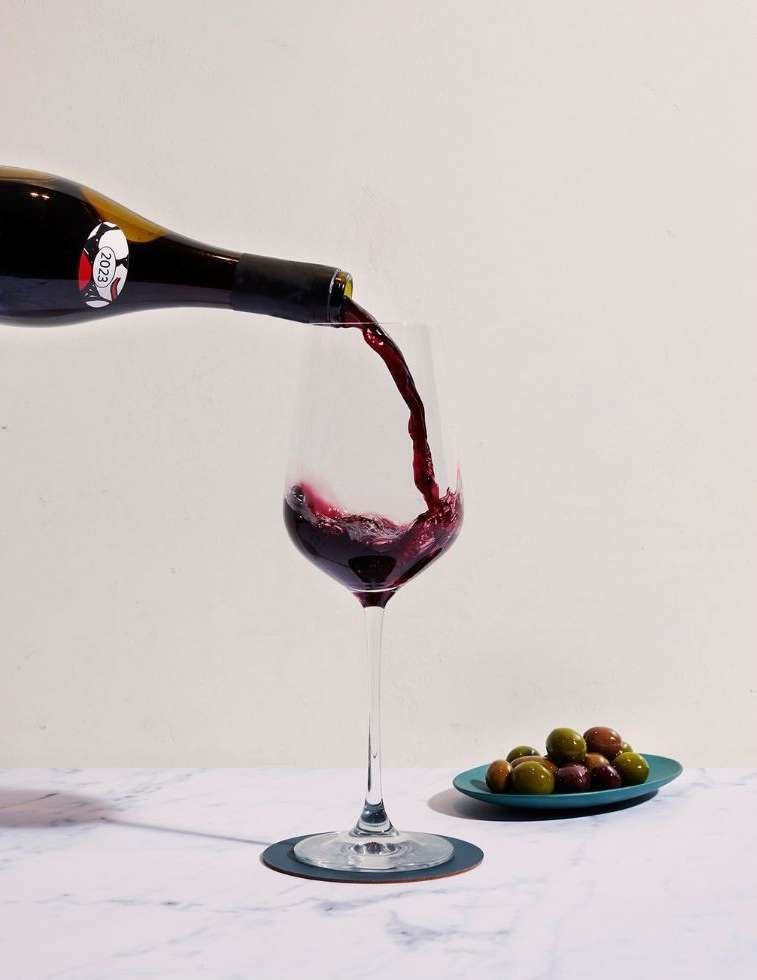 Red wine pours into a stemmed wineglass on a marble surface, with a plate of green and brown olives visible against a white background.