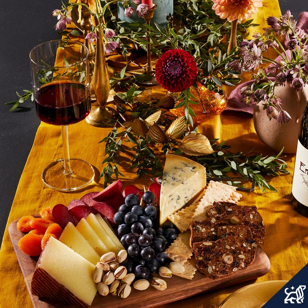 Overhead shot of charcuterie board with cheese, fruit, nuts, crackers, and wine, set on a gold tablecloth with flowers and candles.