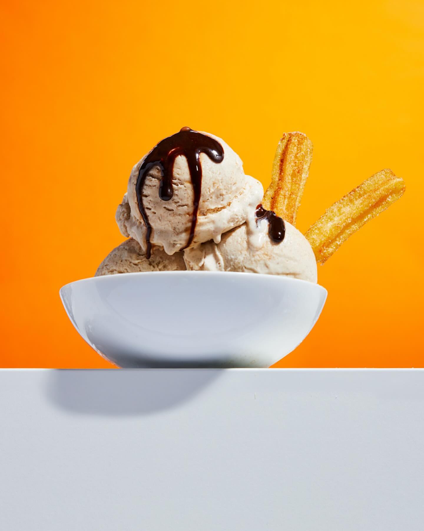 Close-up of a white bowl filled with scoops of ice cream, drizzled with chocolate sauce, and accompanied by churros, against orange backdrop.