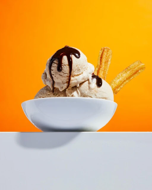 Close-up of a white bowl filled with scoops of ice cream, drizzled with chocolate sauce, and accompanied by churros, against orange backdrop.