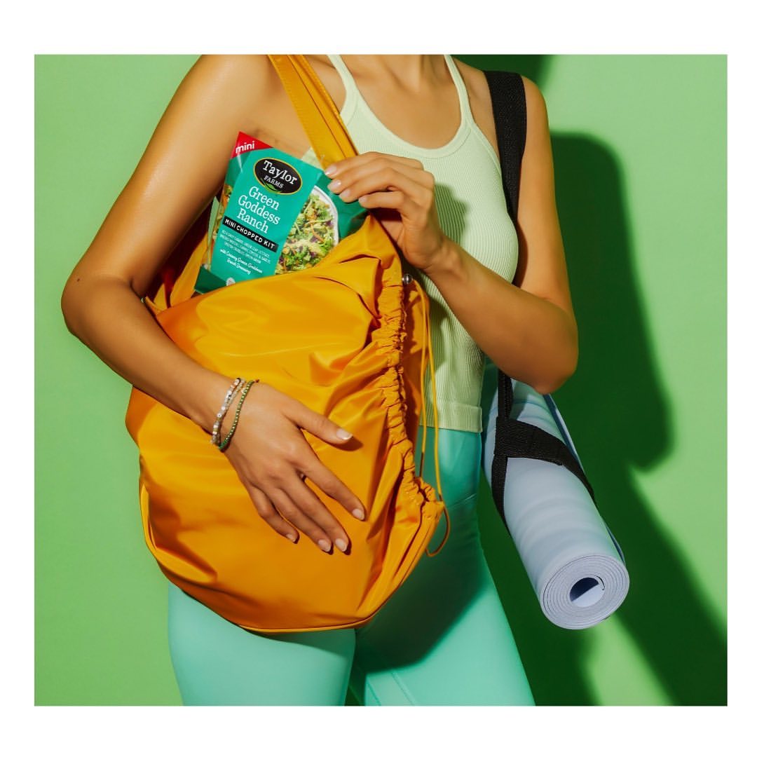 A person with a yellow drawstring bag on their shoulder, containing salad and a yoga mat against a green background.