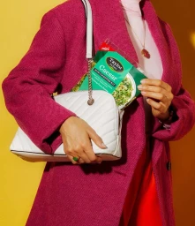 Woman in pink textured coat holding a white quilted handbag containing a bagged salad kit against a yellow background.