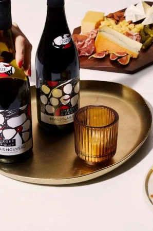 Two wine bottles and a ribbed amber candle holder on a brass tray beside a charcuterie board with cheese and figs.