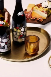 Two wine bottles and a ribbed amber candle holder on a brass tray beside a charcuterie board with cheese and figs.