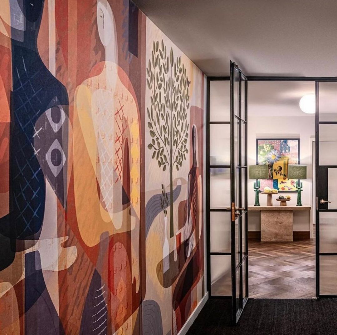 Hallway interior featuring a colourful mural with figures and a view into a room with table lamps and a herringbone floor pattern.