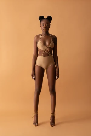 Black woman wearing a beige cutout swimsuit and gold heels against a solid warm tan studio background
