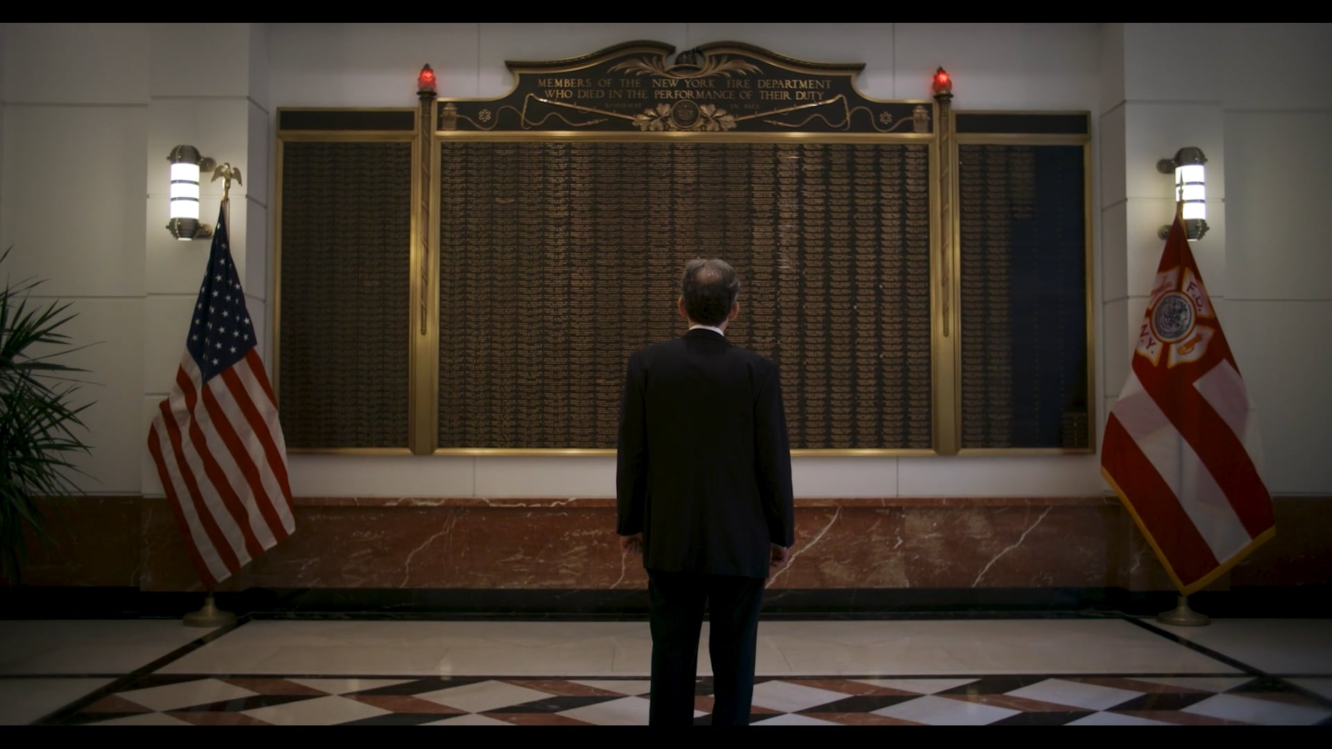 A man in a dark suit stands facing a large memorial plaque flanked by flags and wall sconces inside a grand interior space.